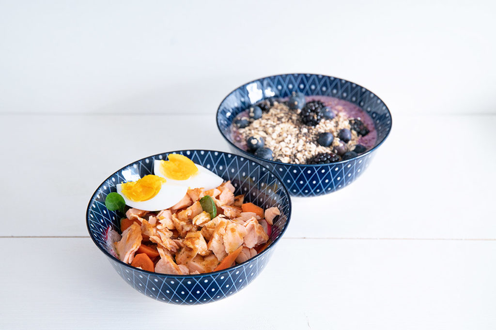 2 bowls nebeneinander: eine gefüllt mit Fisch und Ei, die hintere mit einem Beeren.Smoothie.