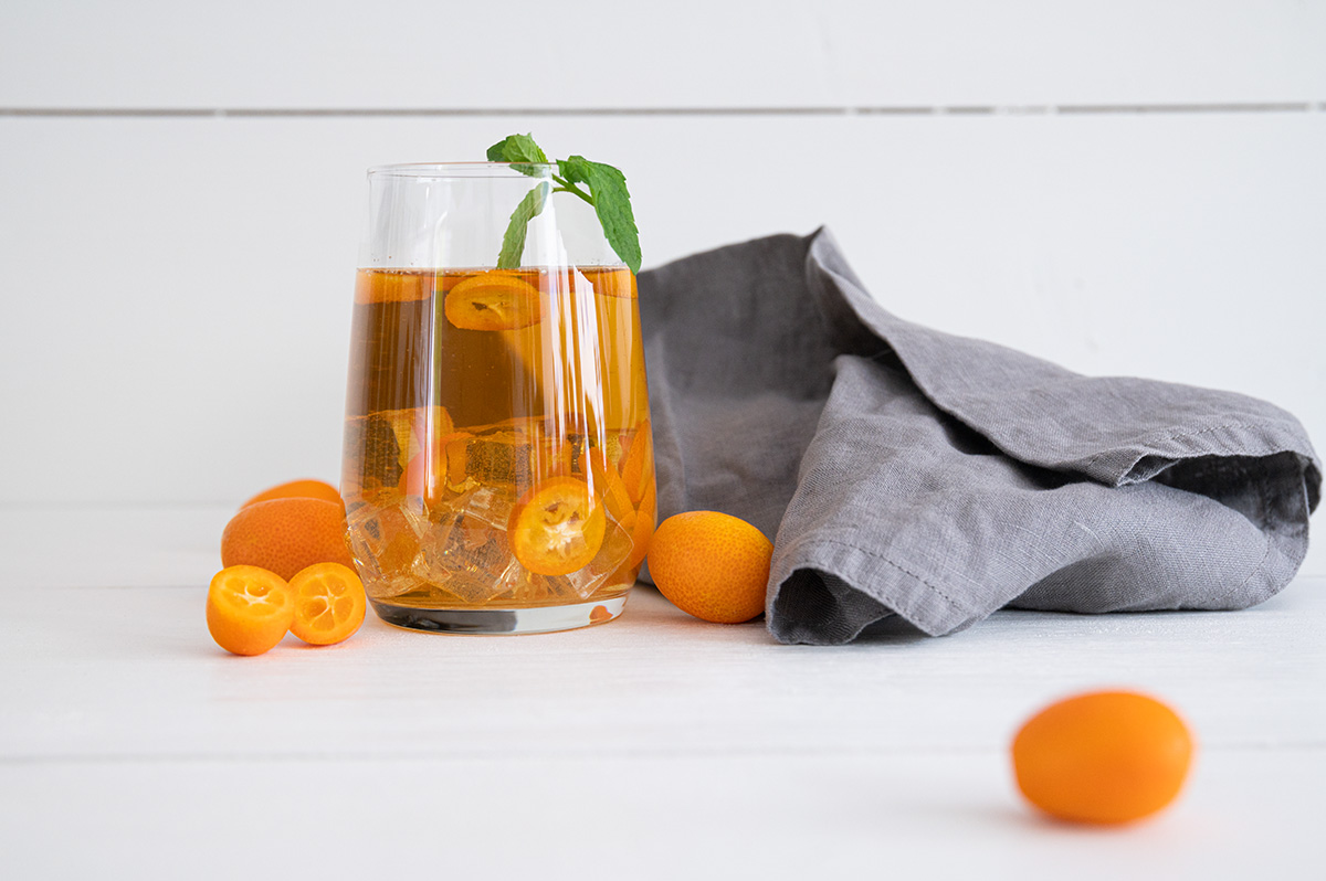 Ein Glas gefüllt mit Eiswürfeln, Kumquathälften und Kumquat-Eistee, daneben noch einige Kumquats die lose darum liegen.