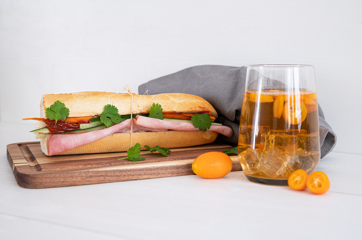 Ein vietnamesisches Sandwich belegt unter anderem mit Gurken, Karotten, Schinken und Koriander auf einem Holzbrett, daneben ein Glas Kumquat Eistee.