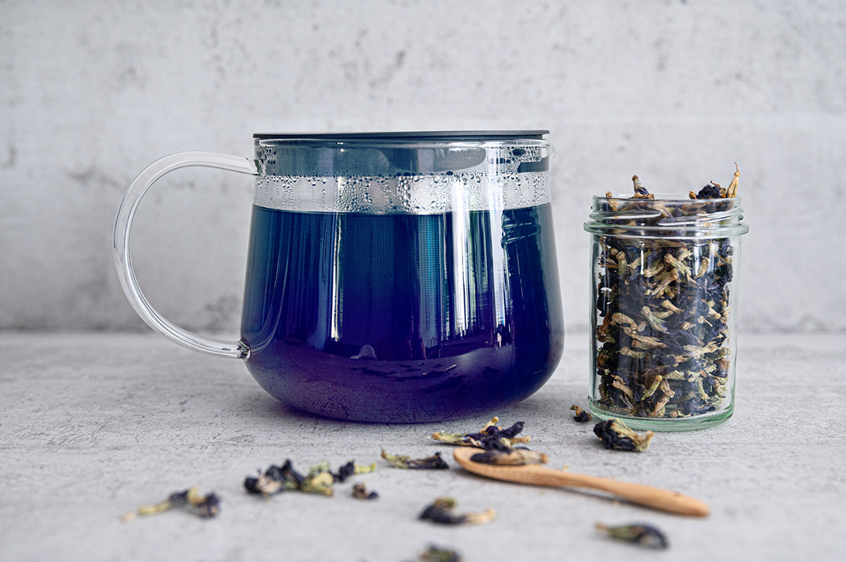 Glaskanne mit blauem Butterfly Pea Tea neben Glas mit getrockneten Butterfly Pea Tea Blüten.