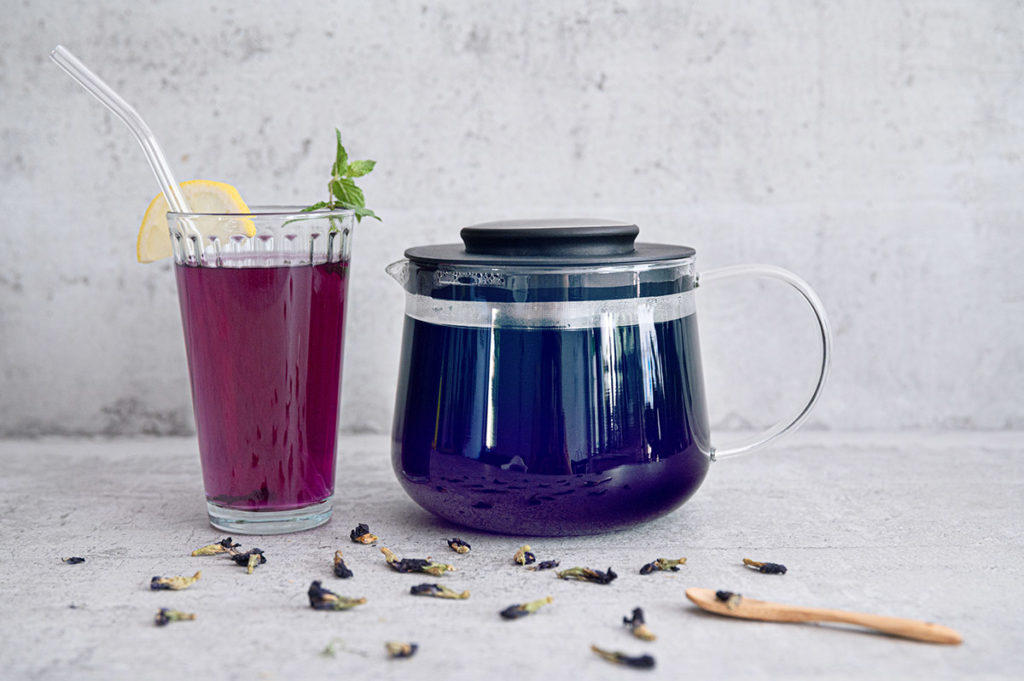 Eine Glaskanne mit blauem Tee neben einem Glas mit lila Tee.