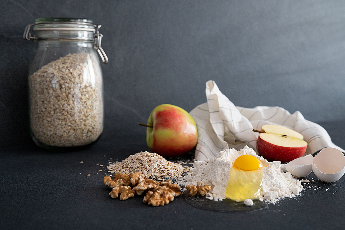 Verschiedene Zutatenfür Apfelbrot wie Mehl, ein bereits aufgeschlagenes Ei. Äpfel und Haferflocken.