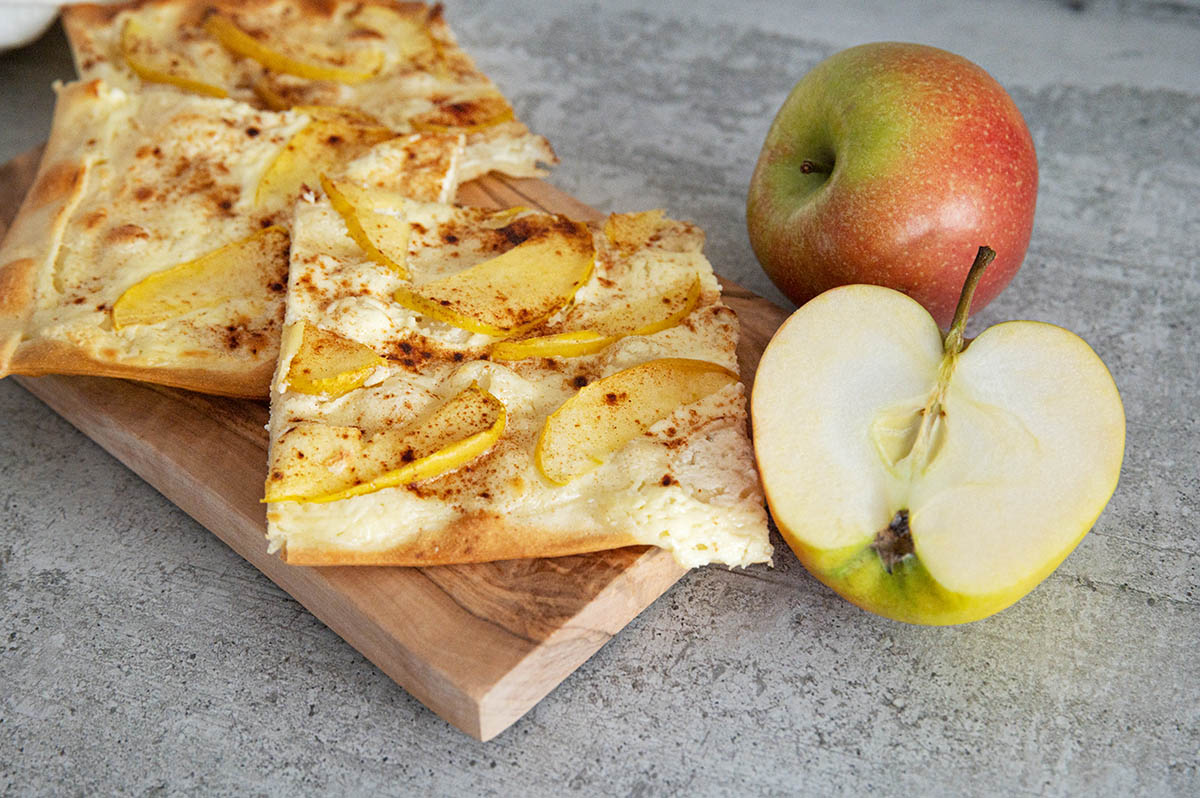 Flammkuchen mit Apfel serviert auf Holzbrett, daneben ein ganzer und ein halber Apfel.