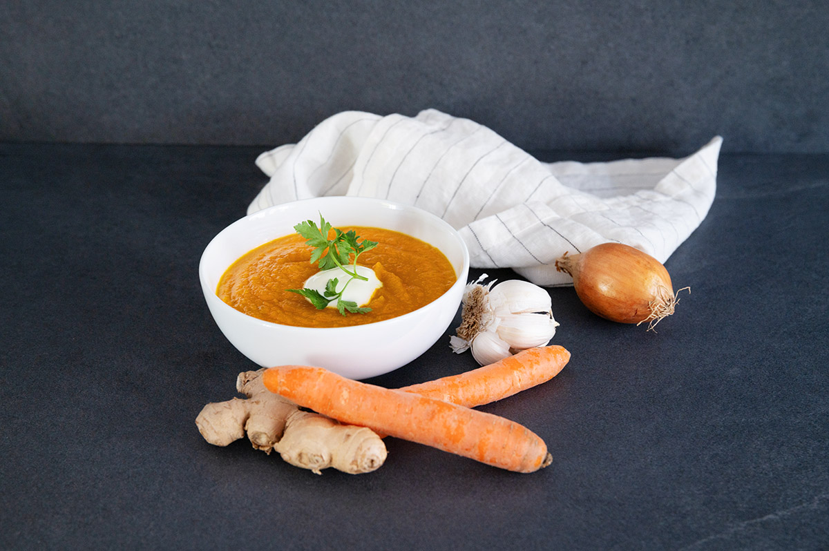 Eine Detox Gemüse Suppe aus Kurkuma und Karotten in einer weißen Suppenschüsse angerichtet, im Vordergrund Karotten, Ingwer, Knoblauch und Zwiebel als Deko.