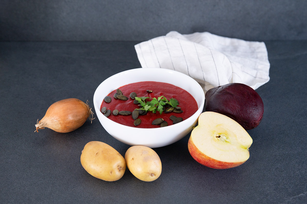 Schüssel Rote Bete-Suppe, daneben Zwiebeln, Kartoffeln, ein halber Apfel und eine Rote Bete.