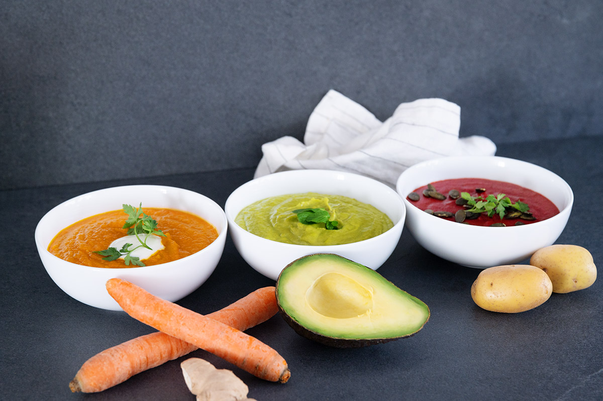 Die drei vorgestellten Detox Gemüse Suppen nebeneinander, sowie Karotten Avocado und Kartoffeln davor.