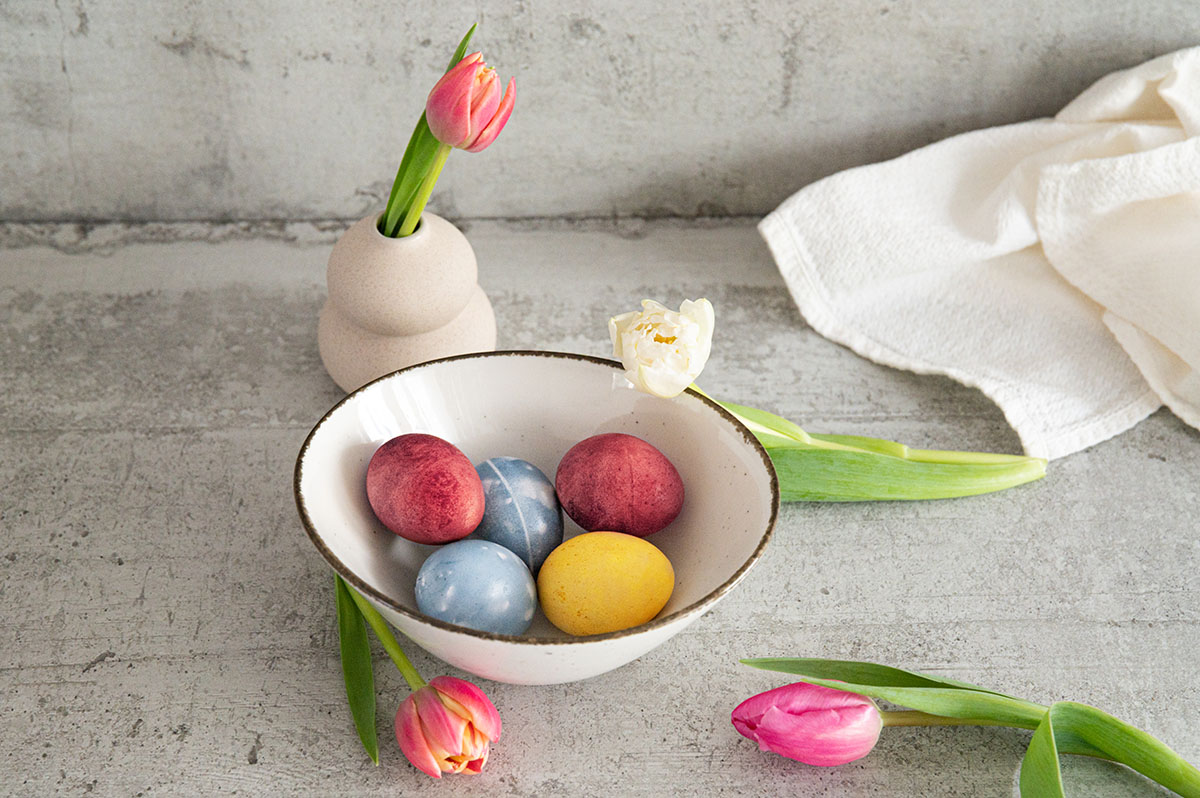 Porzellanschale mit verschhiedenfarbigen Eiern, daneben Tulpen.